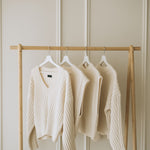 Cream-colored ribbed knitted cardigans hanging on a light wood clothing rack against a beige paneled wall.