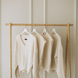 Cream-colored ribbed knitted cardigans hanging on a light wood clothing rack against a beige paneled wall.
